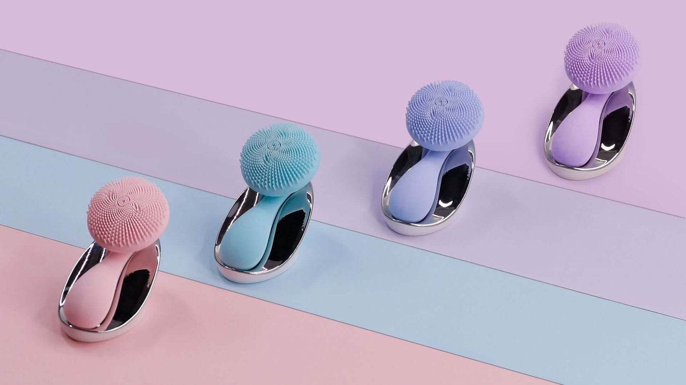 Electric face washer in various colors: pink, blue, and purple against a pastel background